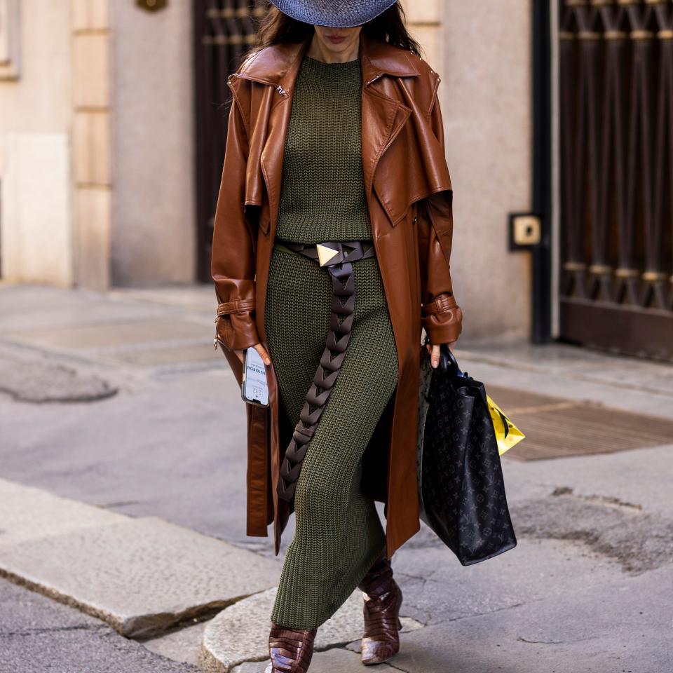 A person in a stylish outfit walks on a sidewalk, wearing a wide-brimmed hat, a long brown leather coat, an olive-green dress, and brown boots, carrying a black bag