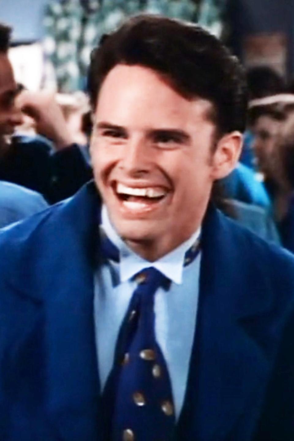 Walton Goggins smiling, wearing a suit with a patterned tie, in a crowded indoor setting