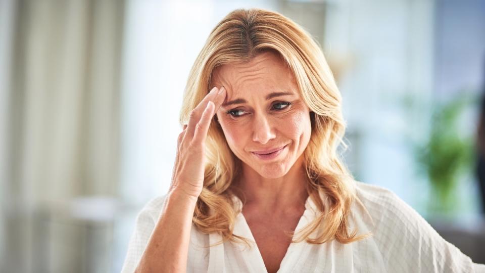 A blonde woman in a while shirt with her hand on her head suffering from menopause brain fog