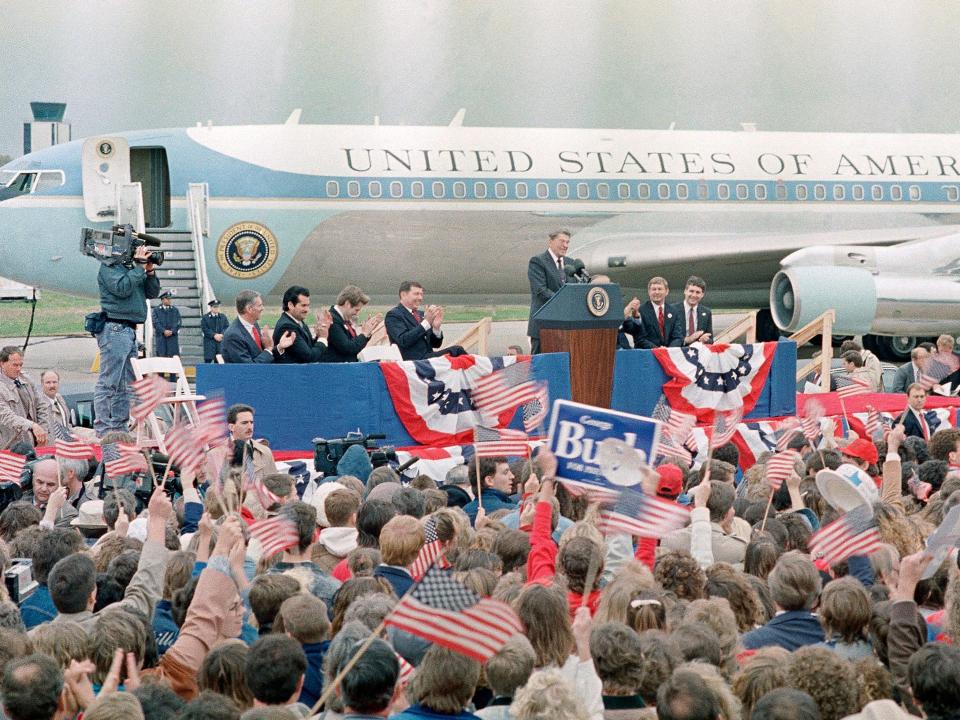 A crowd stands holding American flags faces then President Ronald Reagan, standing behind a podium in front of a blue and white jet