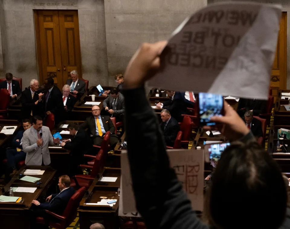 A camera captures the House floor, where a dozen men in suits and ties are in their seats looking disengaged. Digns held by protesters in the gallery.