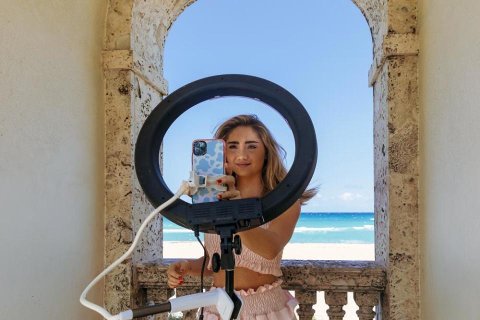 A girl sets up her smartphone on a ring light in front of an archway that looks out onto a bright blue ocean scene.