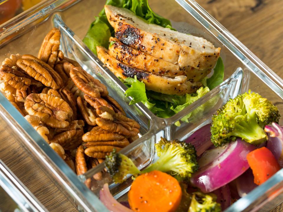 A meal prep container on a wooden table with nuts, chicken, and veggies