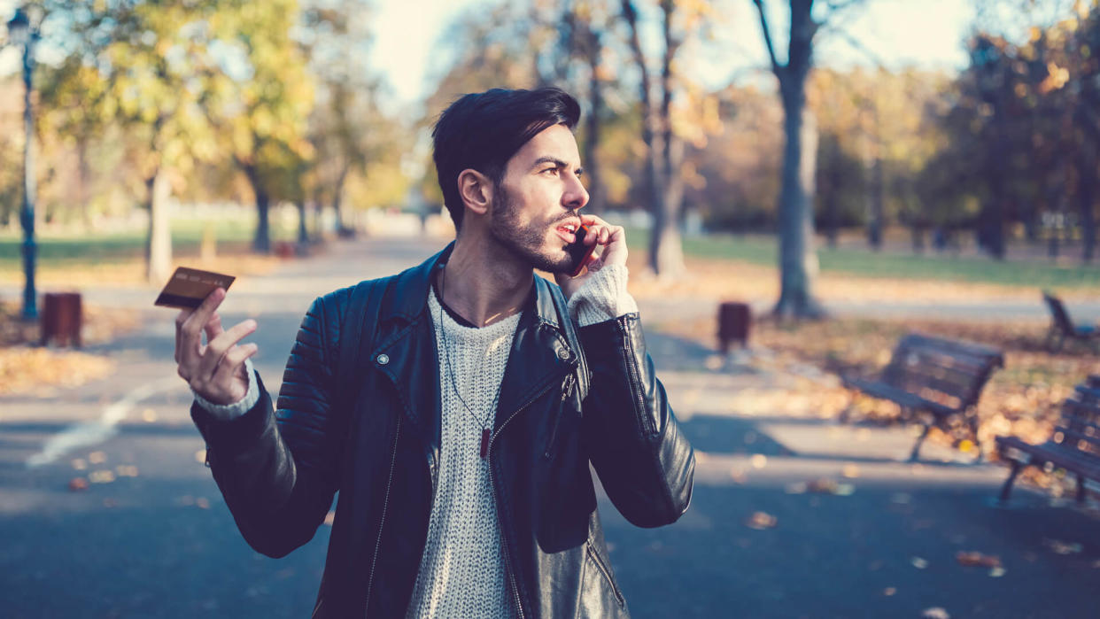 Angry man in the park holding credit card and talking on the phone.