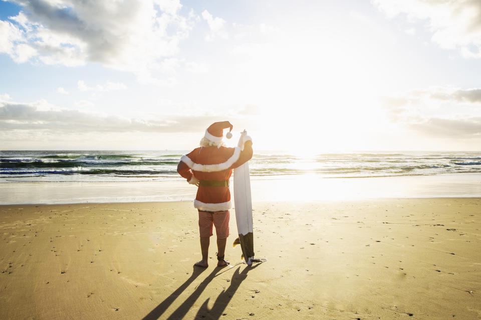 A man dressed as Santa Claus holds a surfboard while looking at the ocean.