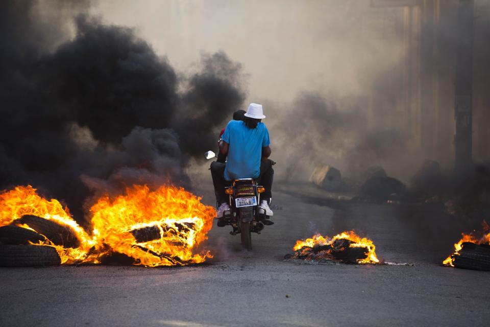 People on a motorcycle drive past a burning roadblock placed by anti-government protesters who are demanding the resignation of Haitian President Jovenel Moise, near the presidential palace in Port-au-Prince, Haiti, Wednesday, Feb. 13, 2019. Protesters are angry about skyrocketing inflation and the government's failure to prosecute embezzlement from a multi-billion Venezuelan program that sent discounted oil to Haiti. (AP Photo/Dieu Nalio Chery)