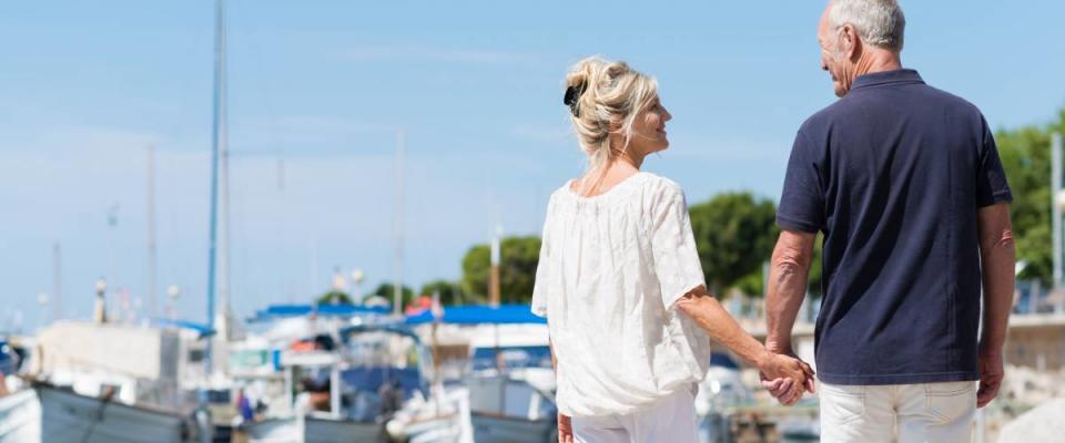 Mature couple enjoying a day at the coast walking away from the camera hand in hand past a small boat harbour