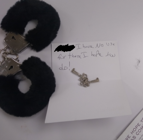 Handcuffs with fake fur and a note reading "I have no use for these I hope you do!" on a white surface