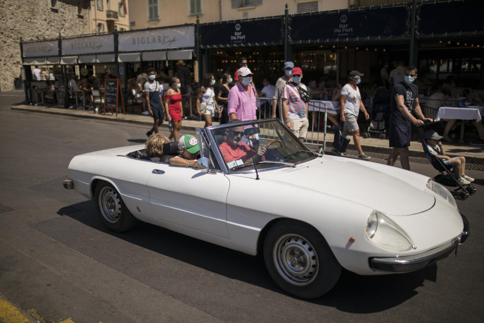 Tourists wearing face masks look on as a classic sports car drives through the Saint-Tropez port in southern France, Saturday Aug 8, 2020. The glamorous French Riviera resort of Saint-Tropez is requiring face masks outdoors starting Saturday, threatening to sober the mood in a place renowned for high-end, free-wheeling summer beach parties. (AP Photo/Daniel Cole)