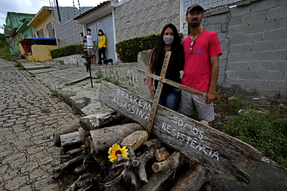 Paulo Sergio Doe, his wife Alessandra Cavalcante Doe and their children pictured in the background, stand in front of their home, in the Pinheiro neighborhood of Maceio, Alagoas state, Brazil, Sunday, March 6, 2022. The couple is standing behind a wooden cross marker with text that reads in Portuguese; "Braskem Assassin. We are Resistance," as they refuse to leave their home because of the threat of ground subsidence and geological problems caused by the Braskem mine, that has caused the evacuation of more than 55 thousand people in the city. (AP Photo/Eraldo Peres)