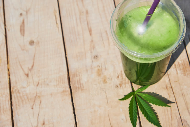 A cannabis leaf lying on a wooden surface next to a glass containing a green liquid.