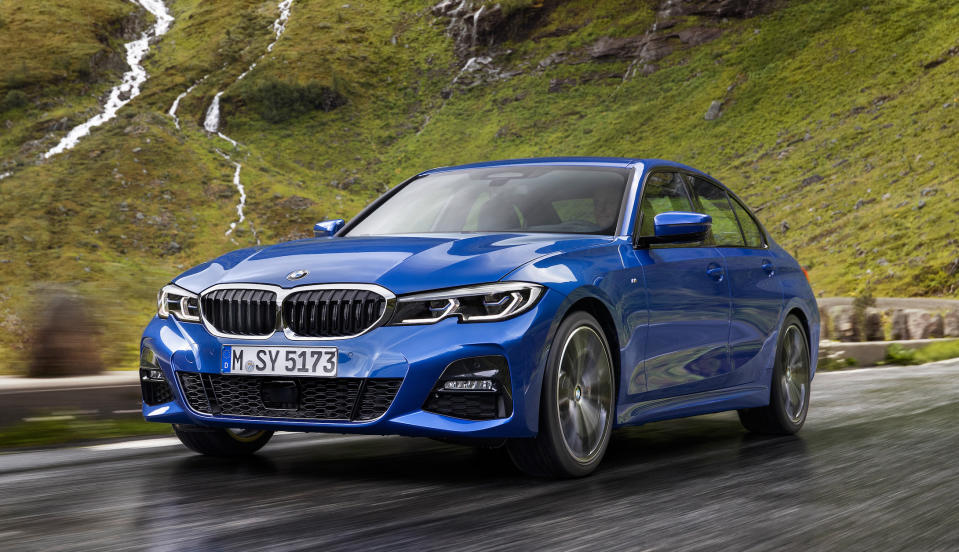 A blue 2019 BMW 3 Series, a compact luxury sedan, driving on a road with a grassy mountain in the background