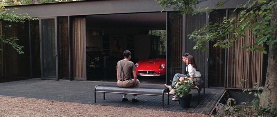 three teenagers sit on benches outside a home garage with a bright red ferrari inside. the home is a designed house in highland park, with glass doors that open into the showroom garage