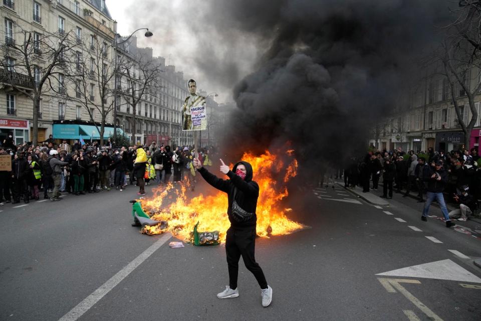 A demonstrator holds a poster mocking French President Emmanuel Macron next to burning garbage cans during a demonstration on Tuesday, March 28, 2023 in Paris (AP)