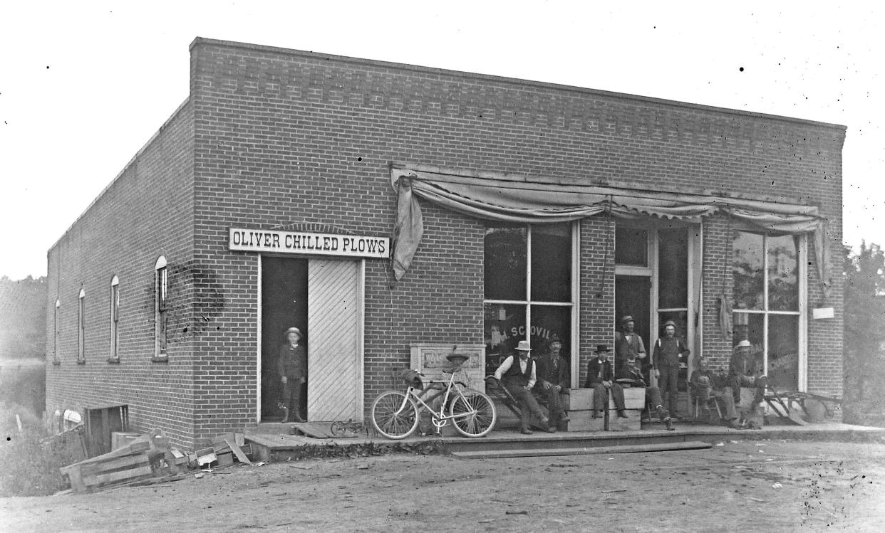 Gentlemen relax outside a brick building circa 1900. A name painted on a window appears to read A.H. Scovill. The building looks rather new. Note the advertisement for plows. Is it a hardware store?