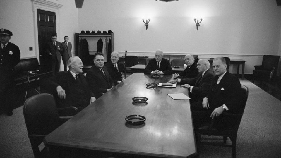 seven men in suits sit around a large table