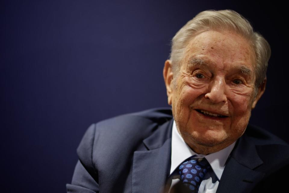 George Soros is seen at an event, smiling. He wears a suit and tie.