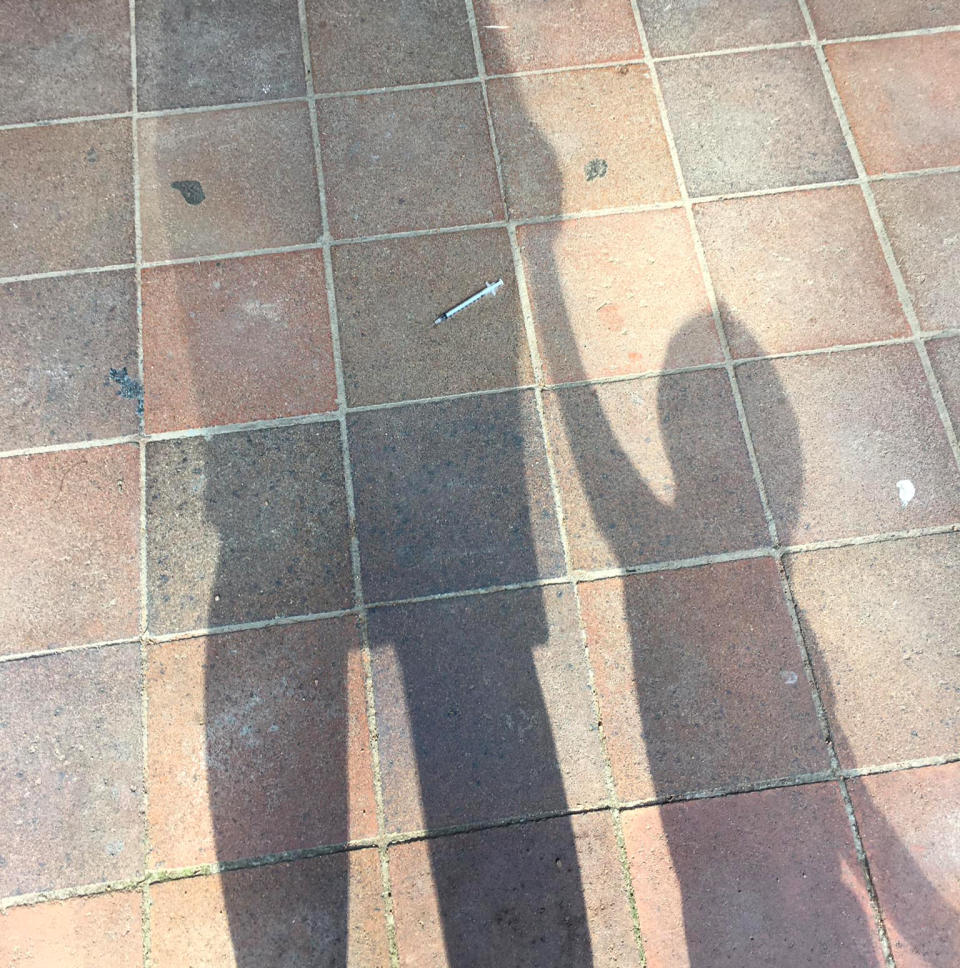 A needle lies on the pavement in front of the shadow of a mum and her child.