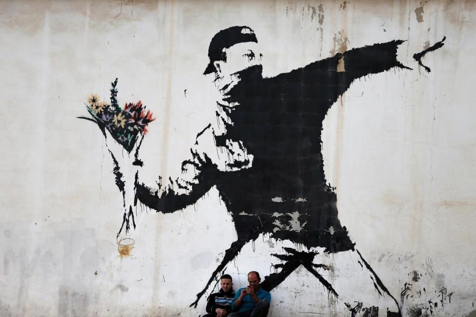 Two men are sitting in front of a famous graffiti of British street artist Banksy, painted on a wall of a gas station in the West Bank city of Bethlehem (Getty Images)