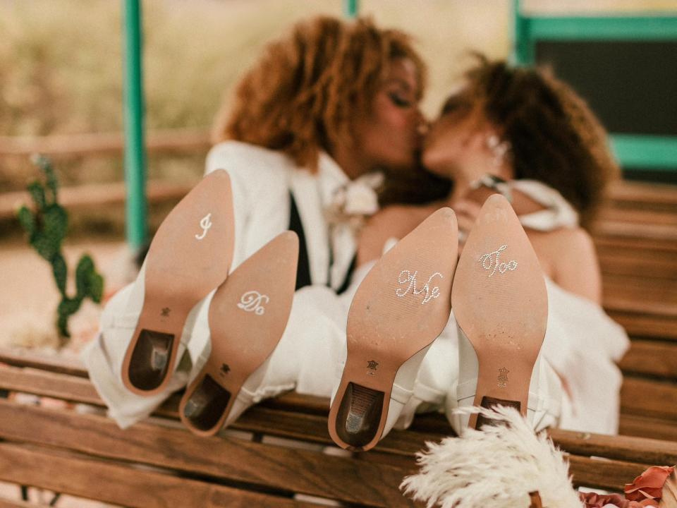 Two brides kiss and show off the bottom of their shoes which say "I Do" and "Me Too."