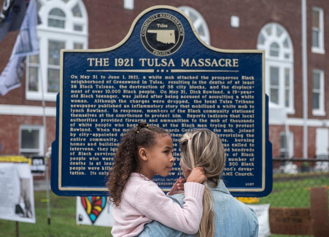 A mother holds her daughter in front of a sign reading: The 1921 Tulsa Massacre.