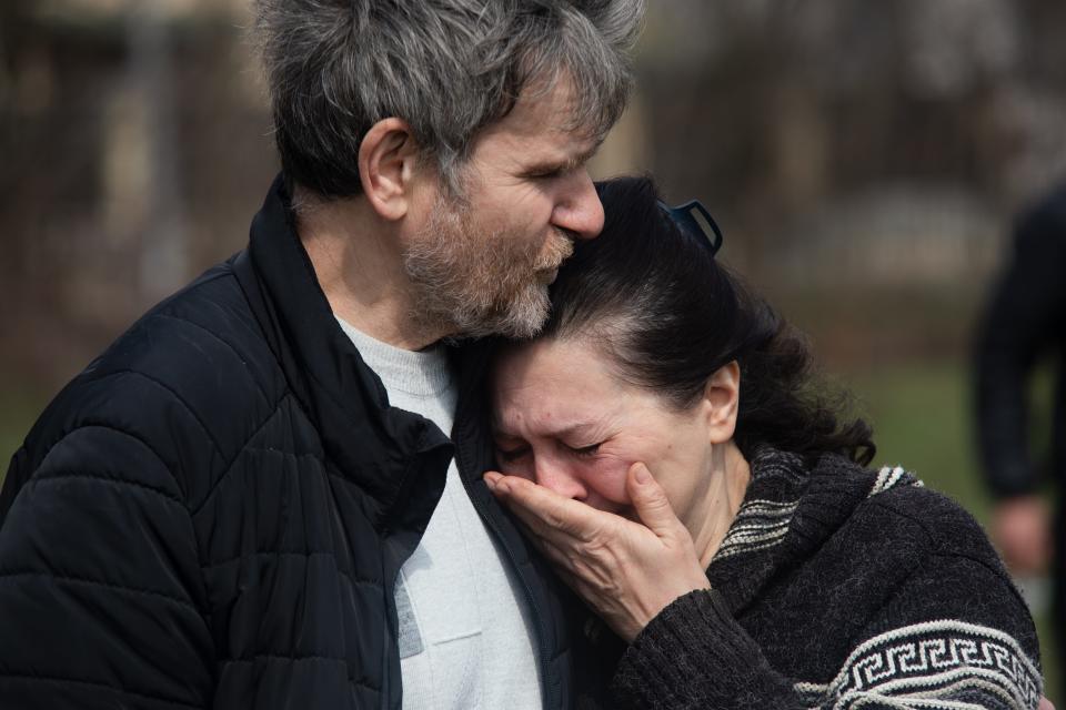 A woman leans against a man as she cries and covers her mouth with her hand.