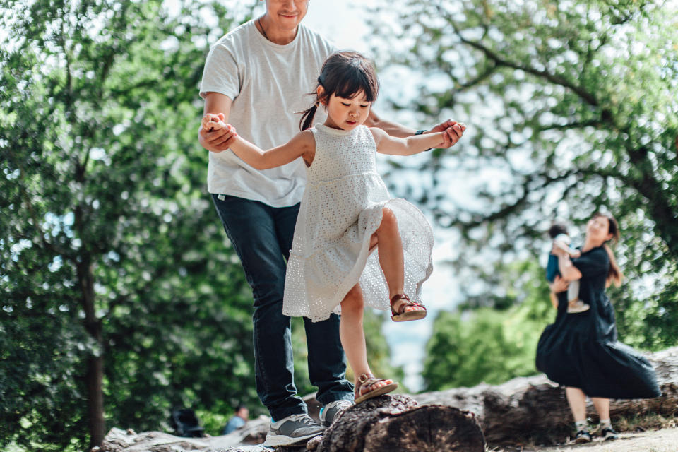 Caring young Asian father holding hands of his little daughter and assisting her to walk along a tree trunk outdoor on a sunny day. Reconnect with nature. Family bonding moments. Outdoor fun.