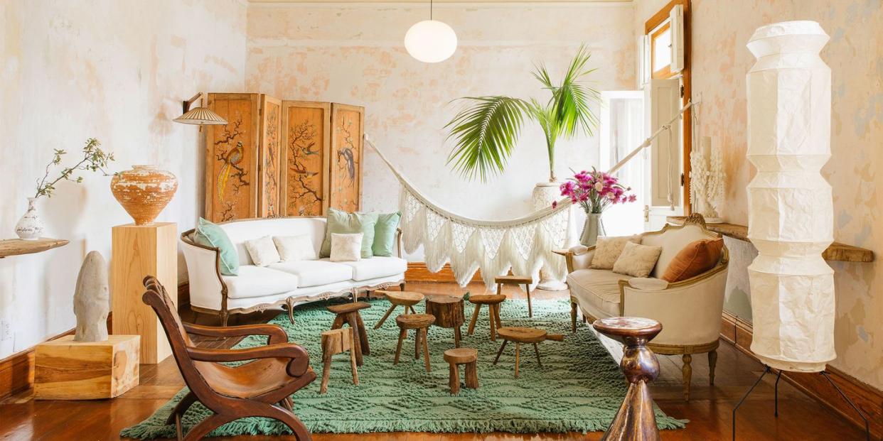 open airy room with a green shaggy rug at center and a series of different shaped wood stools  at center and a white sofa at an angle on the far left and a settee on the right behind which is a tall piece of white column art and opposite are a tall wooden pedestal and a short one with pieces of art on them and a fringed hammock is hung between the two walls at the back behind the furniture