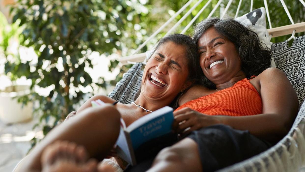 Woman laughing and lying down with daughter in hammock, reading a book on travel. 
