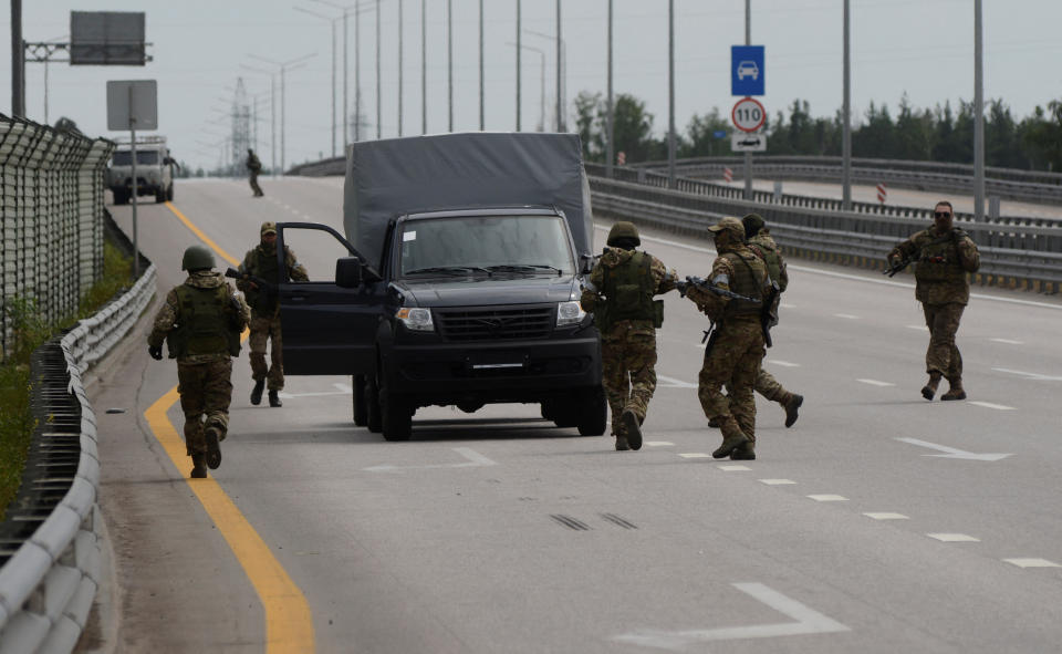 A half dozen military members stand on a large road near a vehicle with an open door.