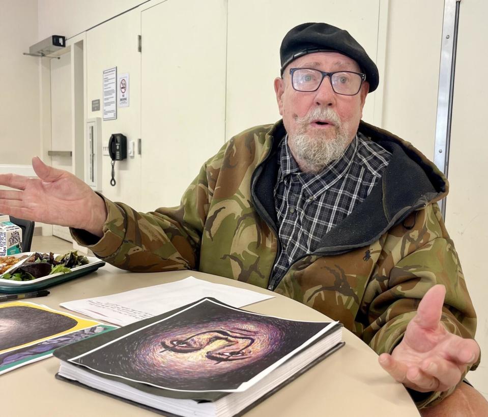 A man in a beret and camouflage jacket, seated at a table with books and papers in front of him