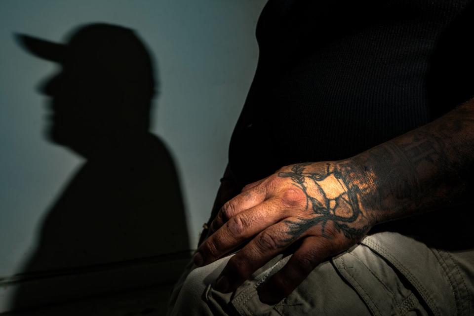 A close up frame of a tattooed hand resting on a person's knee while his shadow is projected on the wall behind.