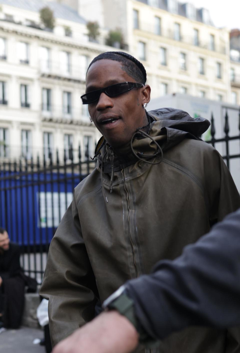 Travis Scott seen outdoors wearing dark sunglasses and a hooded jacket. Urban background with buildings and a fenced area