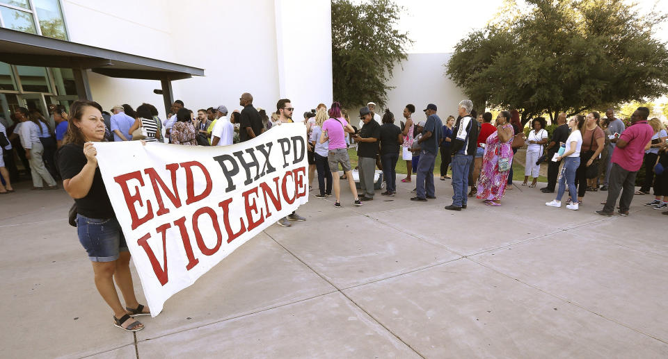 FILE - In this June 18, 2019, file photo, people line up near protesters outside a venue for a community meeting in Phoenix. The meeting stemmed from reaction to a videotaped encounter that surfaced of Phoenix officers pointing guns and yelling obscenities at a black family they suspected of shoplifting. The Phoenix Police Department says it will train officers to track when they point their guns at people. The department announced Friday, Aug. 2 the policy was recommended by the Community Police Trust Initiative and the National Police Foundation, which studied last year's police shootings in the city. The policy will go into effect Aug. 19. (AP Photo/Ross D. Franklin, File)