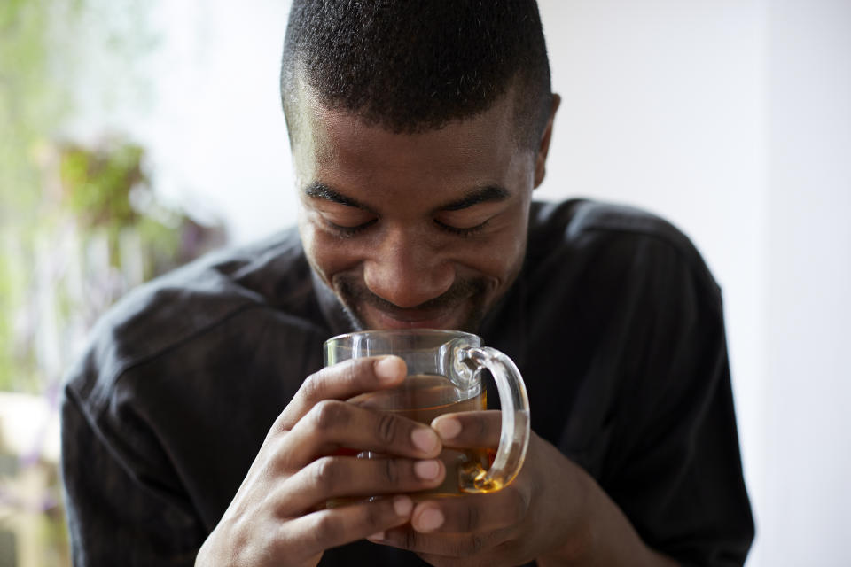 A Black man smiles as he holds a glass cup of tea in his hands up to his face