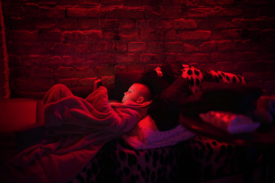 A boy dimly lit in red light lies propped on a pillow on the ground looking at his electronic device.