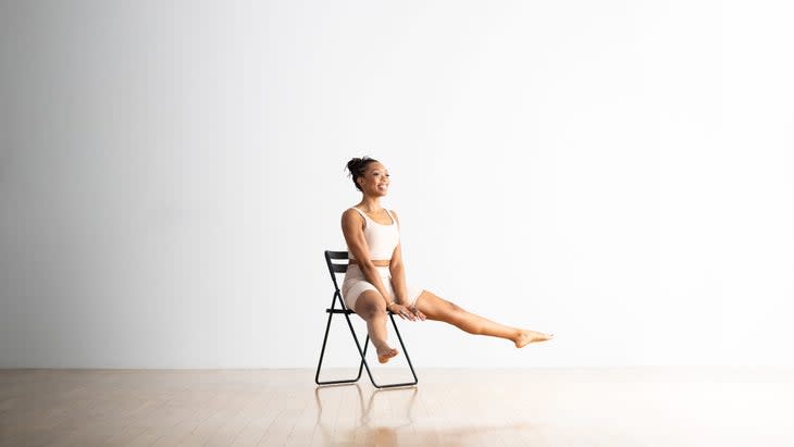 A Black woman sits on a chair practicing Firefly/Tittibhasana. Her legs are lifted and wide and her hands are pressed into the chair seat. The chair is black, the floor is light wood, the background is white