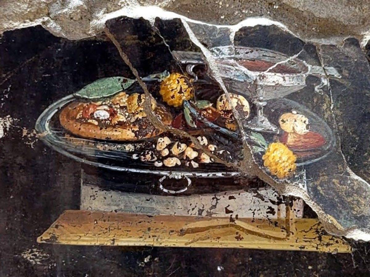 A close up of the fresco found in Pompeii shows a chalice, various fruits, and a circular bread topped with ingredients that ressembles a pizza.