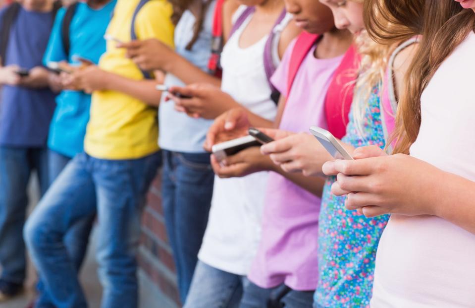 A group of children stand against a wall, all looking down at their smartphones