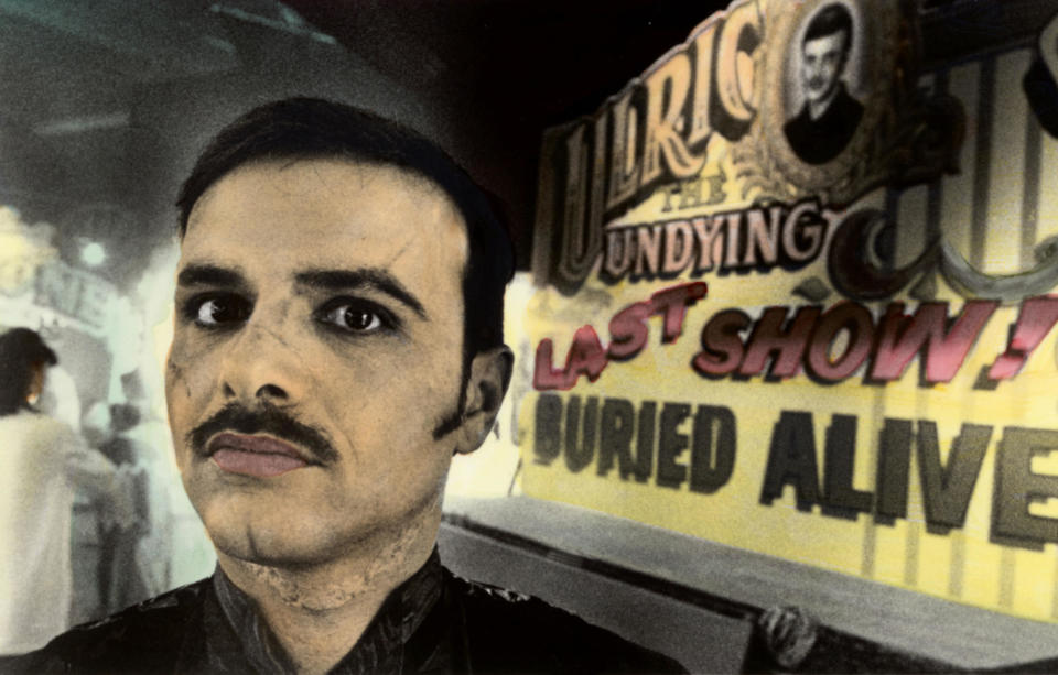 A mustachioed man offers a telling look next to a carnival billboard advertising his living burial