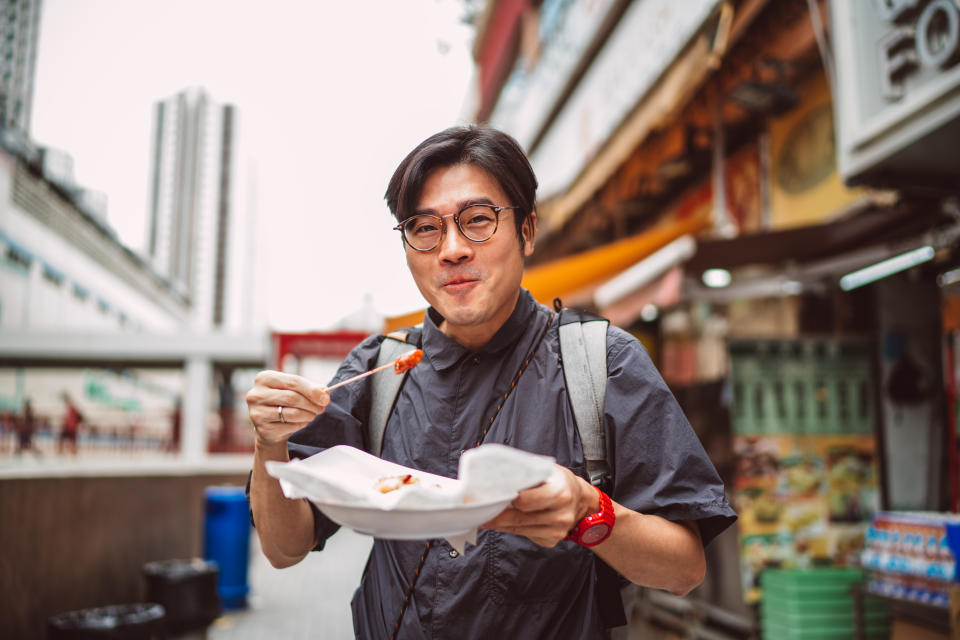 Young handsome man smiling joyfully at the camera while enjoying Hong Kong local street food in street.