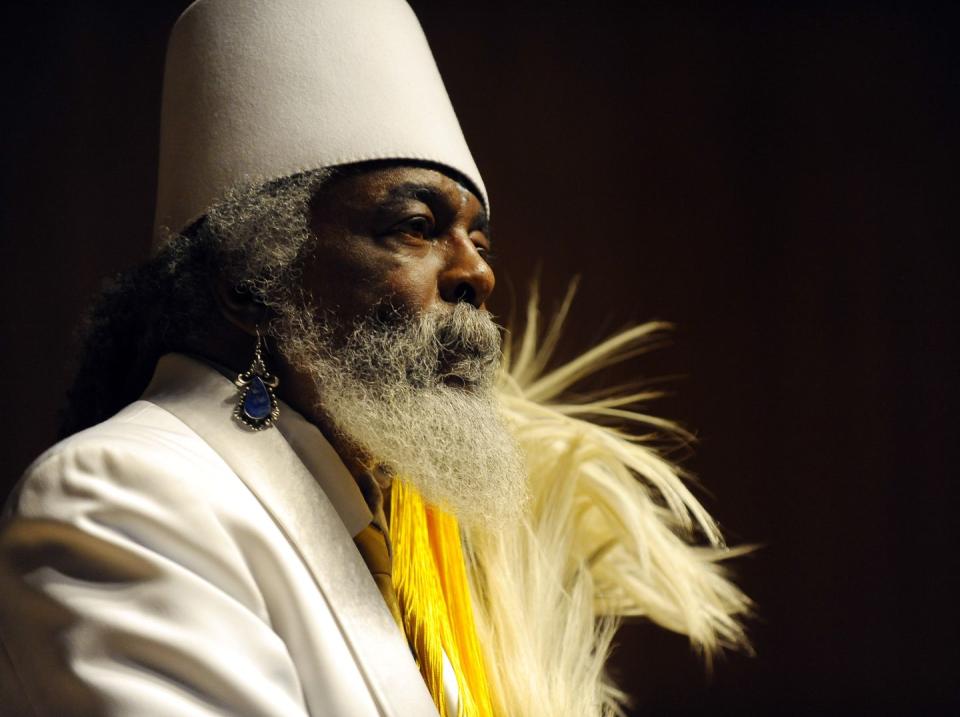 jibreel khazan, wearing a white suit and hat and black earrings, and a bushy gray and white beard, looks off camera