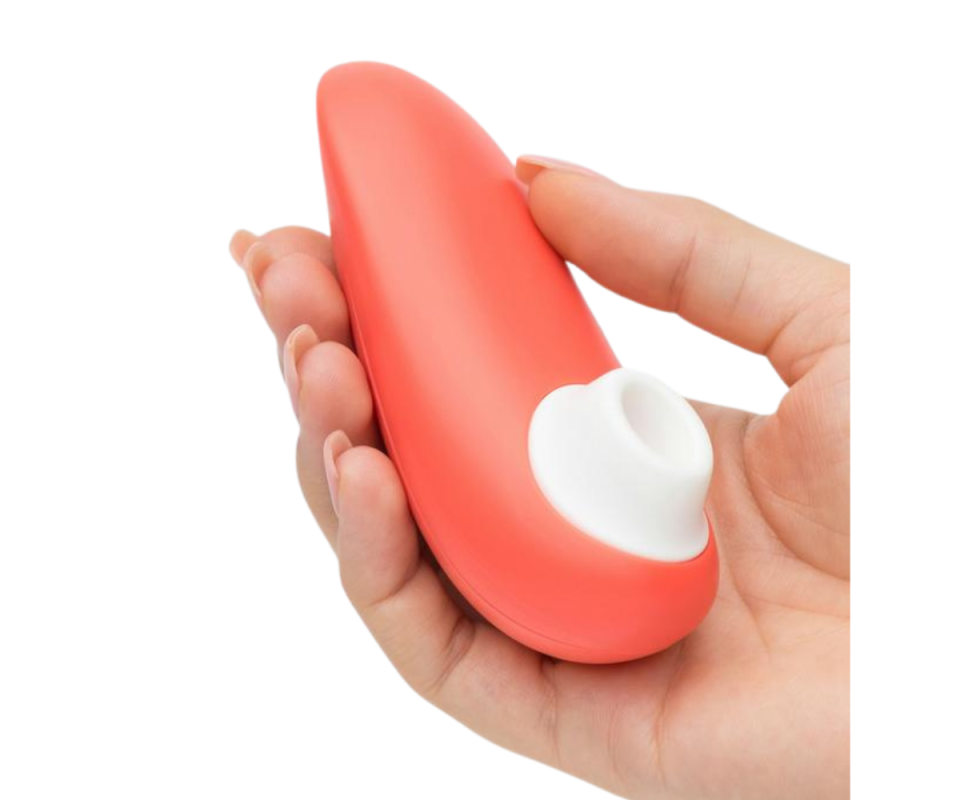 A caucasian woman's hand holds a peach coloured Clitoral Suction Stimulator facing upwards against a white background.