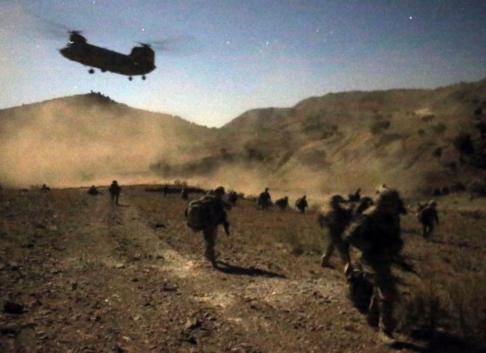 A helicopter flies overhead as soldiers run with their full gear in the desert.