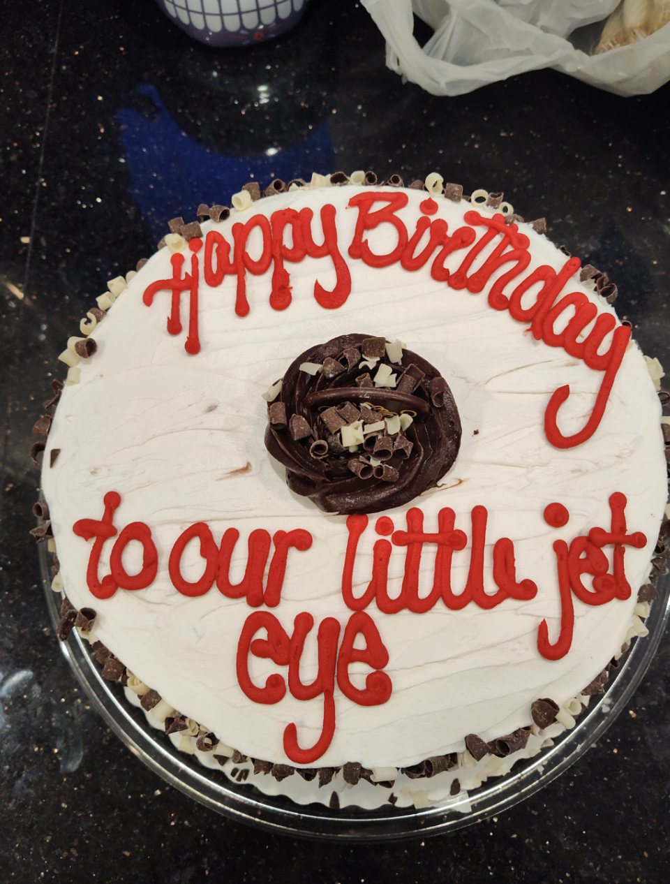 A birthday cake with the message "Happy Birthday to our little jet eye" written in red icing, topped with chocolate pieces