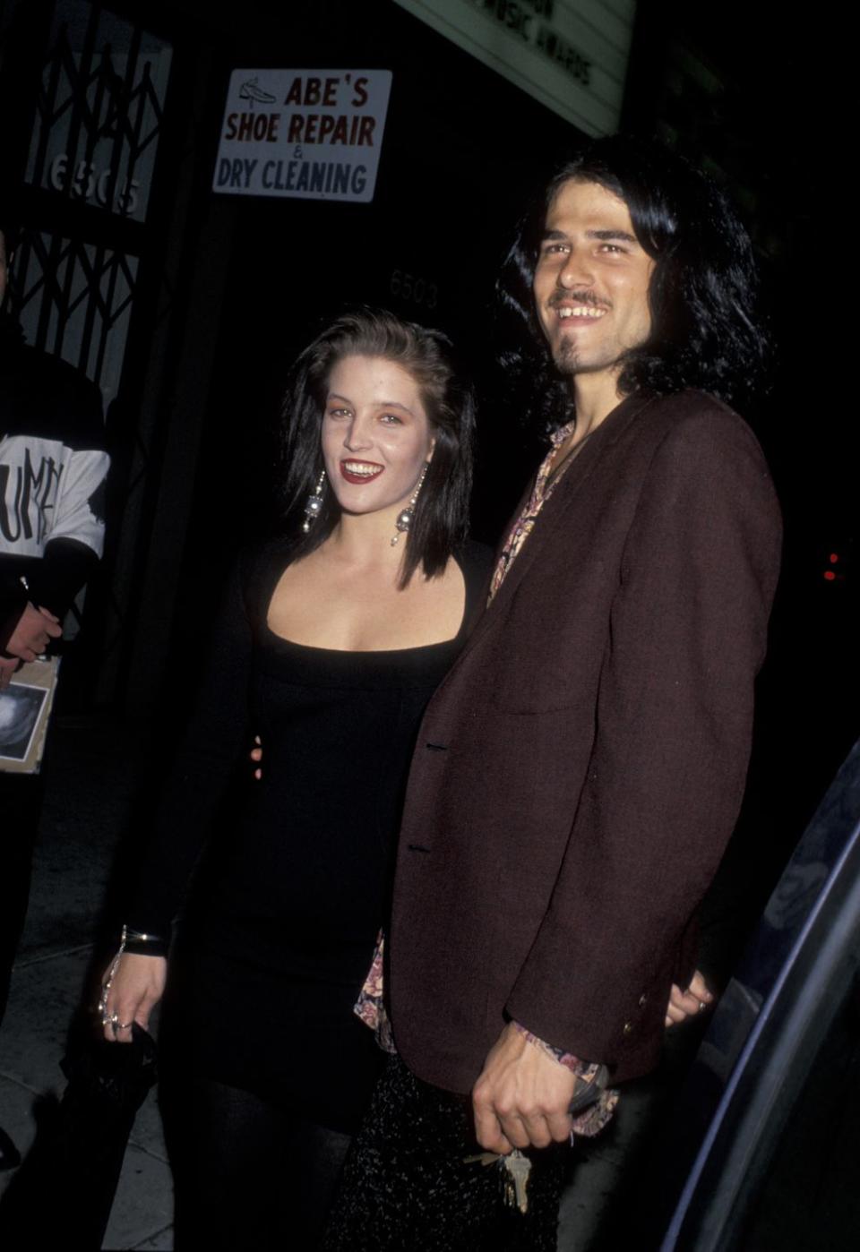 lisa marie presley and danny keough stand next to each other and smile, she wears a black scoop neck dress with long sleeves, he wears a brown suit jacket and holds a set of keys