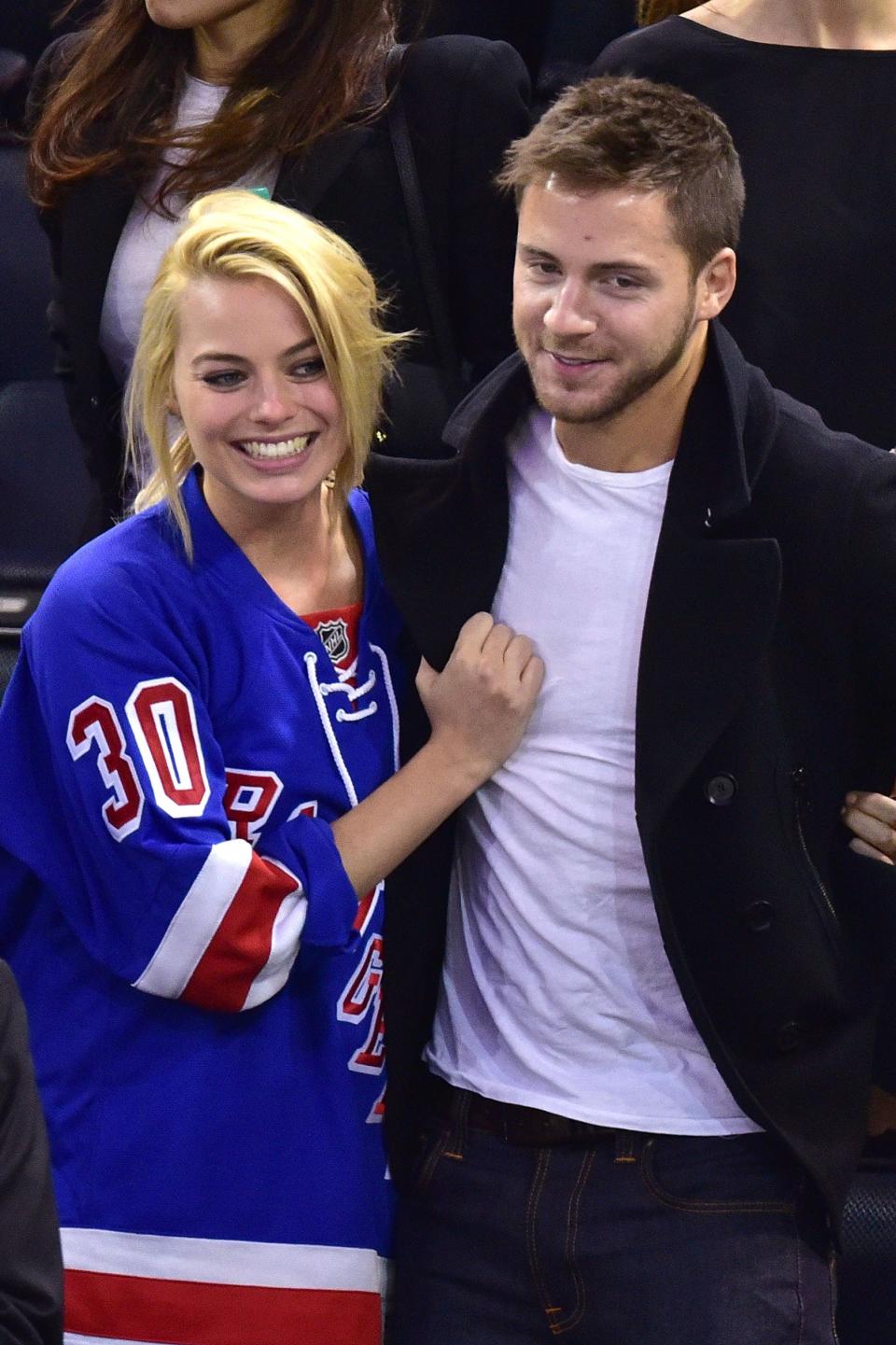 Margot Robbie, wearing a hockey jersey, and Tom Ackerley, in a black coat, smile while standing together at a sports event