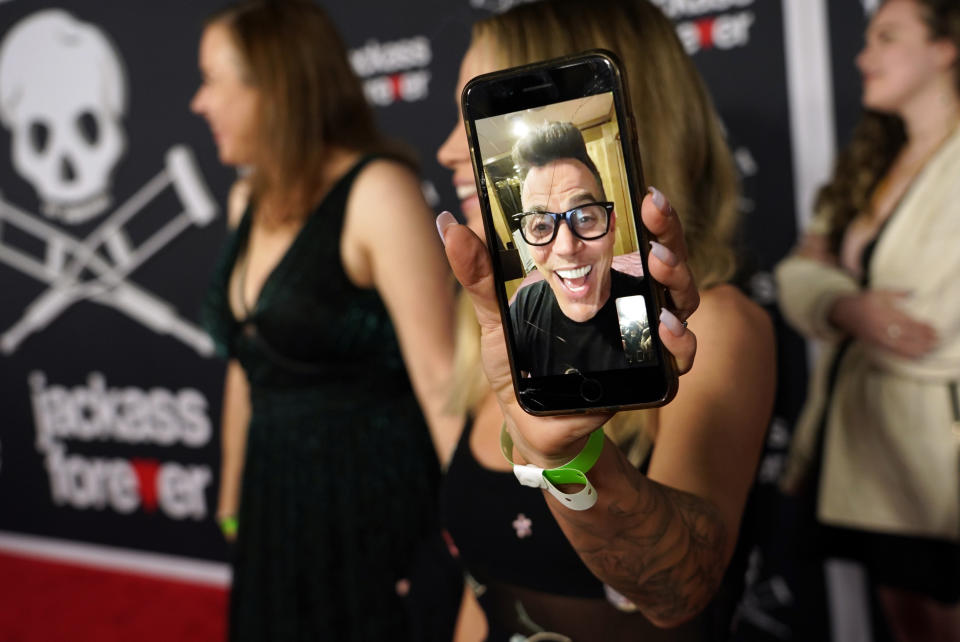 Lux Wright, fiancé of "Jackass Forever" cast member Steve-O, who could not attend the premiere, holds up a live video image of him from her phone on the red carpet, Tuesday, Feb. 1, 2022, at the TCL Chinese Theatre in Los Angeles. (AP Photo/Chris Pizzello)