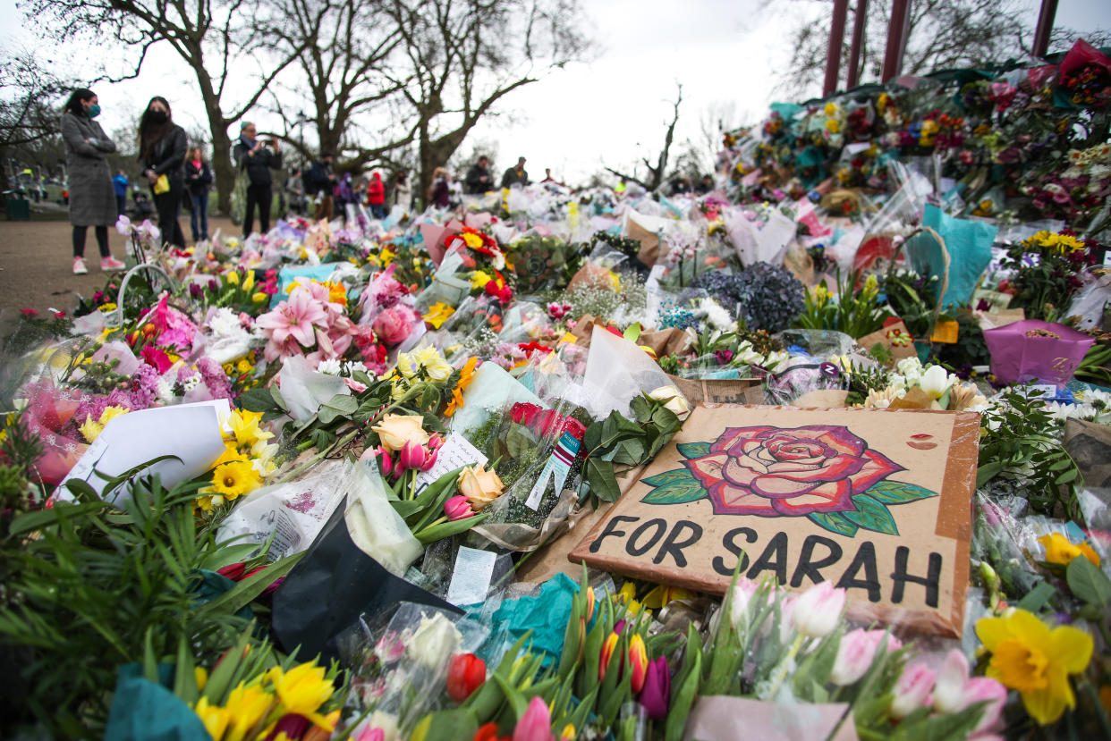 People stand next to floral tributes left at the band stand in Clapham Common, London, for murdered Sarah Everard. Serving police constable Wayne Couzens, 48, appeared in court on Saturday charged with kidnapping and murdering the 33-year-old marketing executive, who went missing while walking home from a friend's flat in south London on March 3. Picture date: Monday March 15, 2021. 