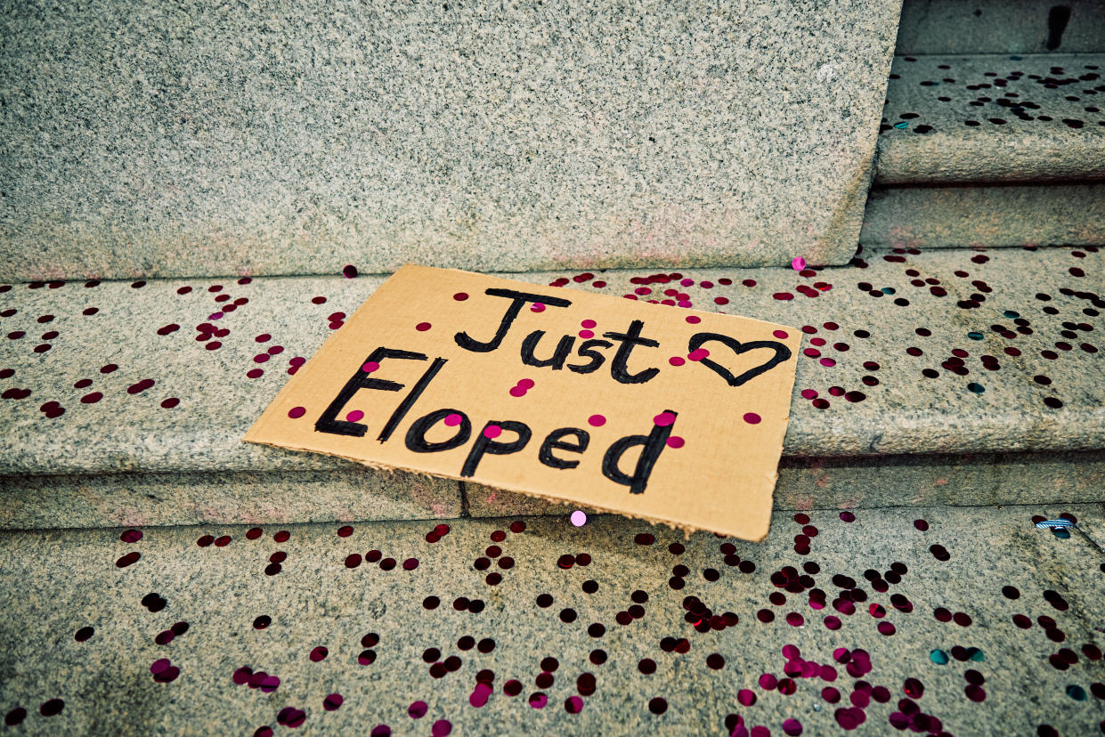 A whimsical cardboard sign reading 'Just Eloped' lies on stone steps, surrounded by a scattering of pink and red confetti, hinting at a spontaneous wedding celebration